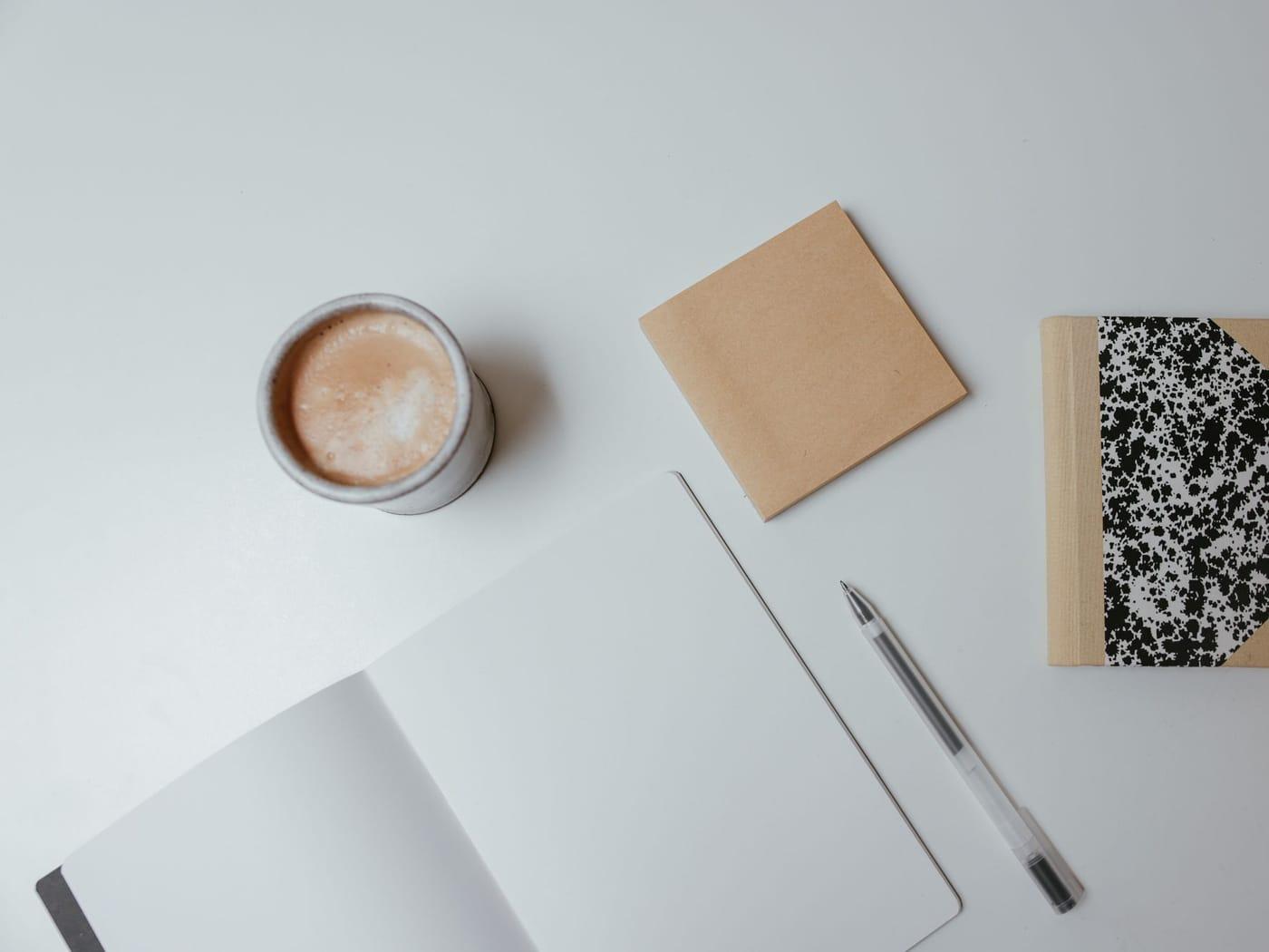 A notebook, pen and cup of coffee laid out on a pale desk
