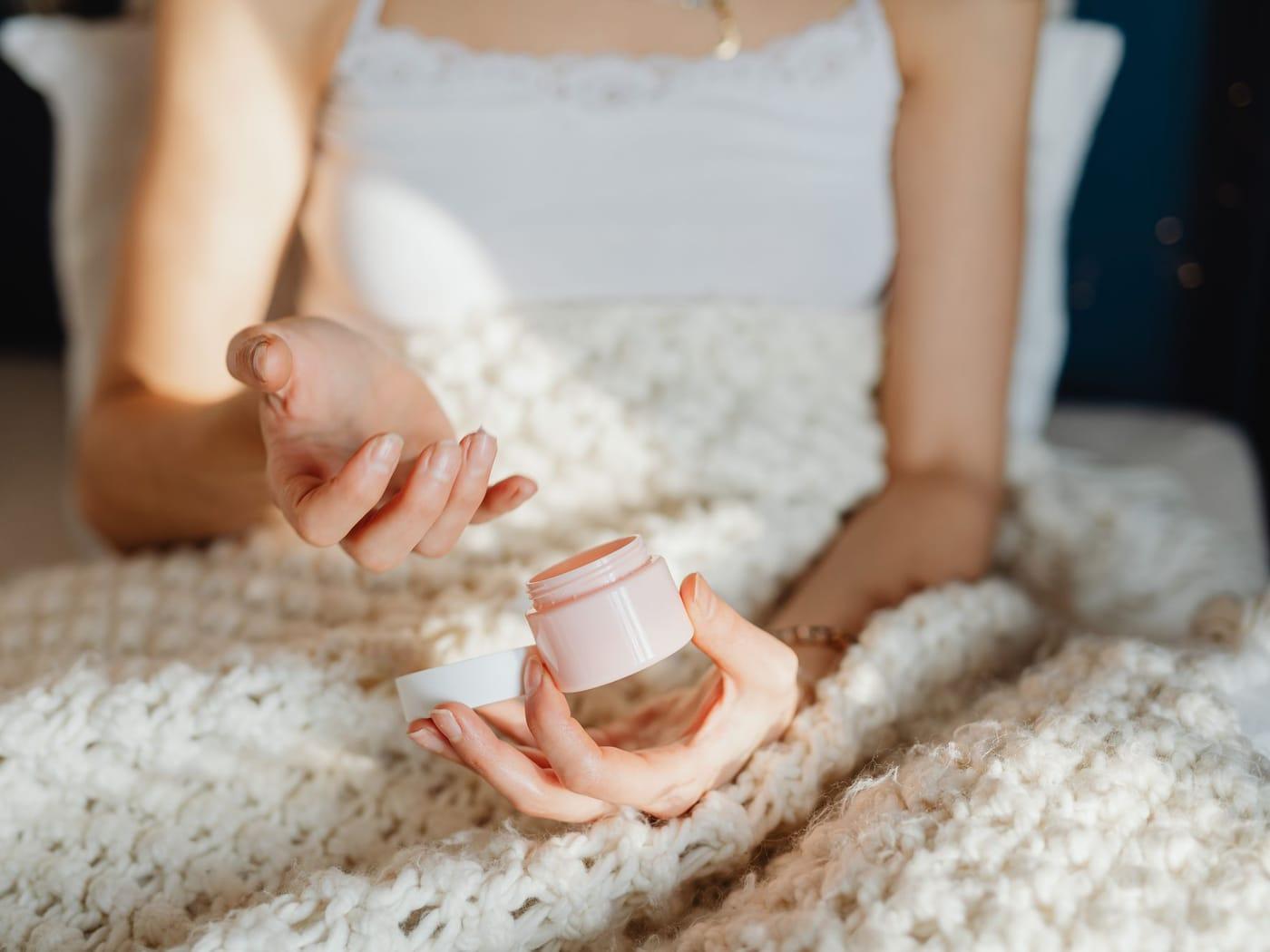 Hands holding an open jar of moisturiser over a chunky knit blanket