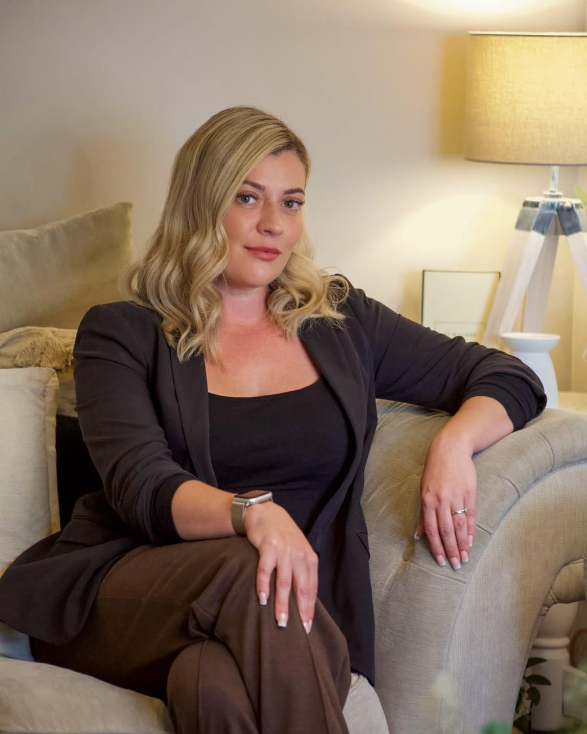 Chelsea Smith in a black blazer, leaning on the arm of a linen sofa beside a softly lit lamp